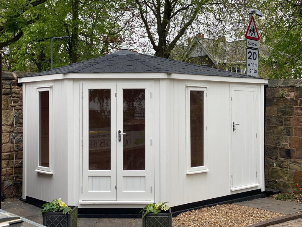 CONTEMPORARY CORNER SUMMERHOUSE PAINTED IN WHITE WITH A BLACK FELT TILED ROOF AND A BUILT IN STORE